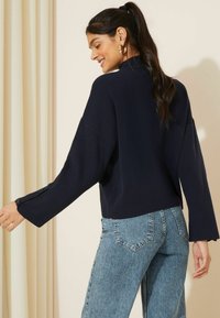 Maglione della marina a maglia con maniche ampie e spacchi laterali, abbinato a jeans azzurri a vita alta. Presenta una texture liscia e un design minimalista.