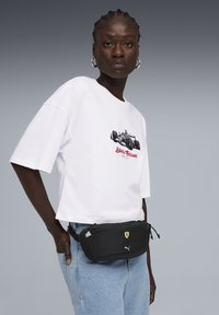Sac banane noir en maille avec les logos Puma et Ferrari, associé à un t-shirt graphique blanc et un jean bleu clair. Design compact avec des fermetures éclair.