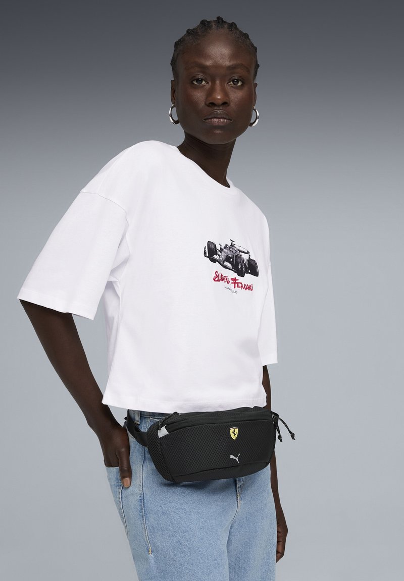 Sac banane noir en maille avec les logos Puma et Ferrari, associé à un t-shirt graphique blanc et un jean bleu clair. Design compact avec des fermetures éclair.