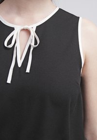 Top senza maniche nero con una rifinitura bianca sul collo, dotato di un dettaglio a chiave con chiusura a fiocco al collo. Tessuto morbido e liscio.