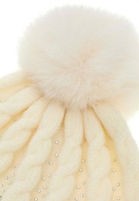 Bonnet en tricot crème avec un pompon duveteux pâle, un design en maille torsadée et de petites embellissements scintillants sur le devant.