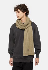Écharpe beige en tissu doux à côtes, avec un patch logo. Portée lâchement autour du cou sur un sweat-shirt gris foncé.