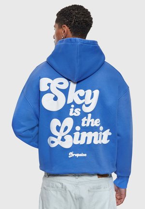 Jeune homme portant un sweat à capuche bleu avec l'inscription "Sky is the Limit" en grandes lettres blanches dans le dos, associé à un pantalon gris clair.