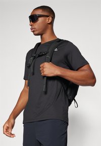 Μαύρο T-shirt και σακίδιο πλάτης, με ρυθμιζόμενους ιμάντες, ματ υφή και μικρό λογότυπο στο στήθος, συνδυασμένο με σκούρα γυαλιά ηλίου.