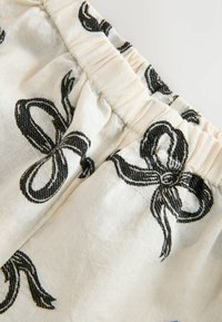 White fabric with black bow print. Gathered elastic waistband with visible stitching. Material appears lightweight and textured.