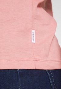Růžové bavlněné tričko s postranními štítky zobrazujícími "Calvin Klein." Tkanina má hladkou texturu a tričko je v kombinaci s modrými džínami.