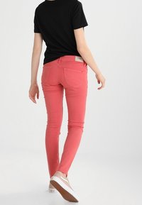 Jeans ajustados en un vibrante color coral, confeccionados en tela de algodón elástico. Presentan bolsillos traseros y pasadores para cinturón. Combinados con una camiseta negra.