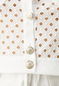 Biała bluzka z teksturowanymi wzorami w kształcie wycięć. Zawiera złote ozdobne guziki oraz usztywniony kołnierz. Wykonana z lekkiego materiału o gładkim wykończeniu.