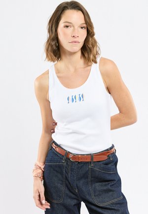 Femme aux cheveux ondulés mi-longs portant un haut blanc sans manches avec un motif de poisson, un pantalon en denim foncé, une ceinture marron et un bandana pêche au poignet.