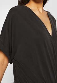 Femme portant un haut noir fluide à col en V et manches courtes, avec un détail de tissu drapé sur le côté, vue des épaules à la taille.
