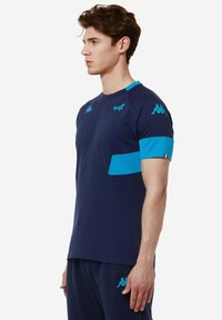 Camisa deportiva de color azul marino con mangas cortas, detalles turquesas en los hombros y en el bolsillo del pecho, textura suave, logo de Kappa en los hombros.