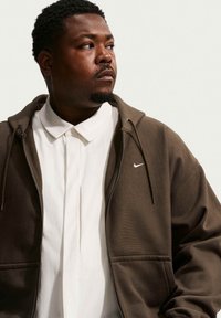 Hombre con cabello corto y rizado, vistiendo una sudadera marrón con cremallera de Nike sobre una camisa blanca con cuello, mirando hacia su izquierda contra un fondo neutro.