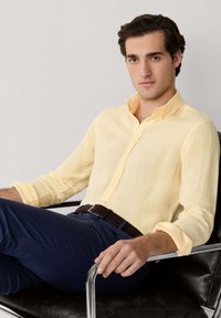 Jeune homme portant une chemise à boutons jaune clair et un pantalon bleu marine, assis avec assurance sur un fauteuil en cuir noir aux accoudoirs métalliques.