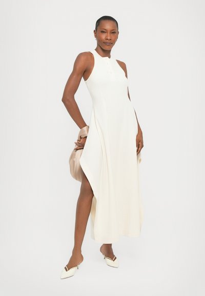 Witte midi-jurk met een mouwloos ontwerp, geribbeld textuur en zijsplitten. Gestyled met een beige tas en witte hiel-slippers.