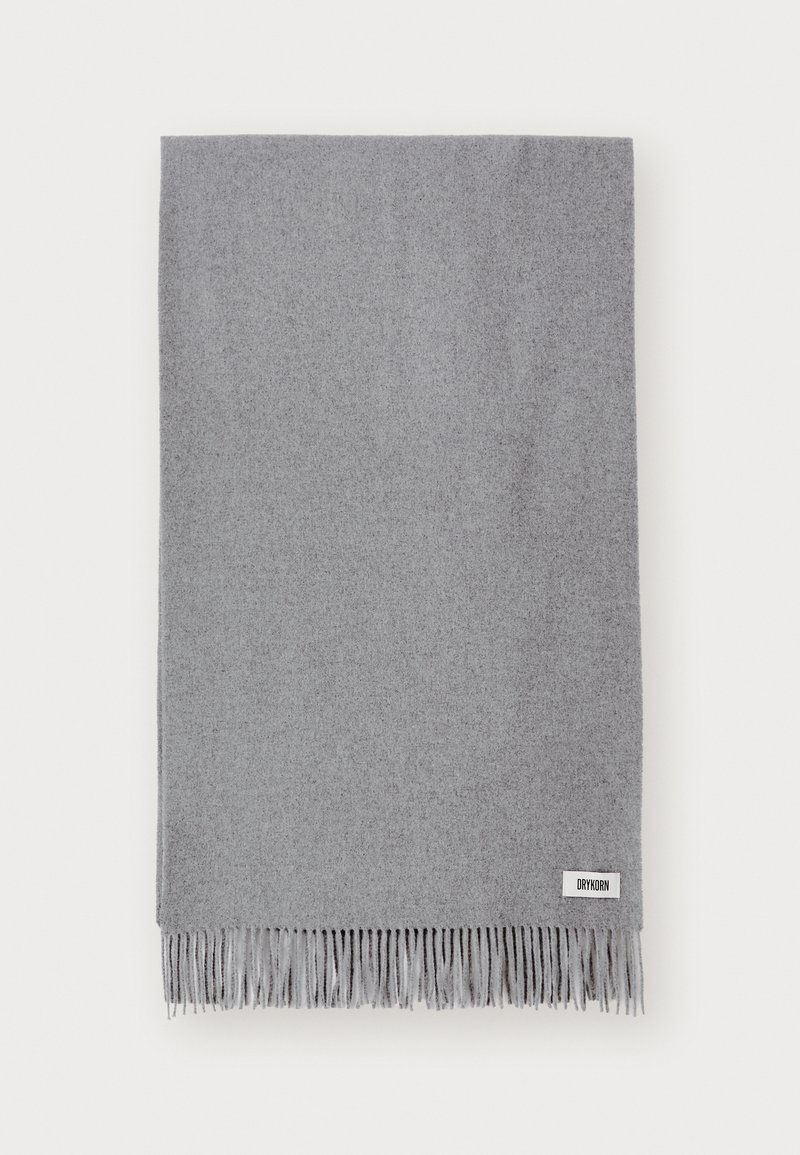 Coperta grigia in lana con bordi frangiati, caratterizzata da una texture morbida e un design minimalista. Include un piccolo etichetta del marchio nell'angolo in basso.