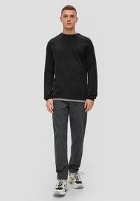 Schwarzes, strukturiertes Langarmshirt kombiniert mit dunkelgrauen Hosen und weißen Sneakers mit schwarzen und grauen Akzenten. Stehende Pose vor einem neutralen Hintergrund.