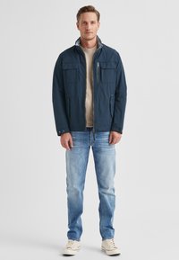 Marineblaue leichte Jacke mit vier Fronttaschen und Reißverschluss. Trägt man über einem beigen Hemd und hellblauen Jeans, kombiniert mit weißen Sneakers.
