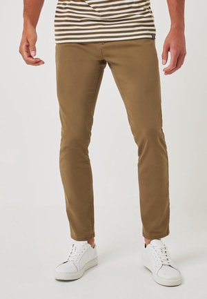 Man draagt nauwsluitende bruine broek, witte sneakers en een beige-wit gestreept shirt, staand tegen een effen achtergrond.