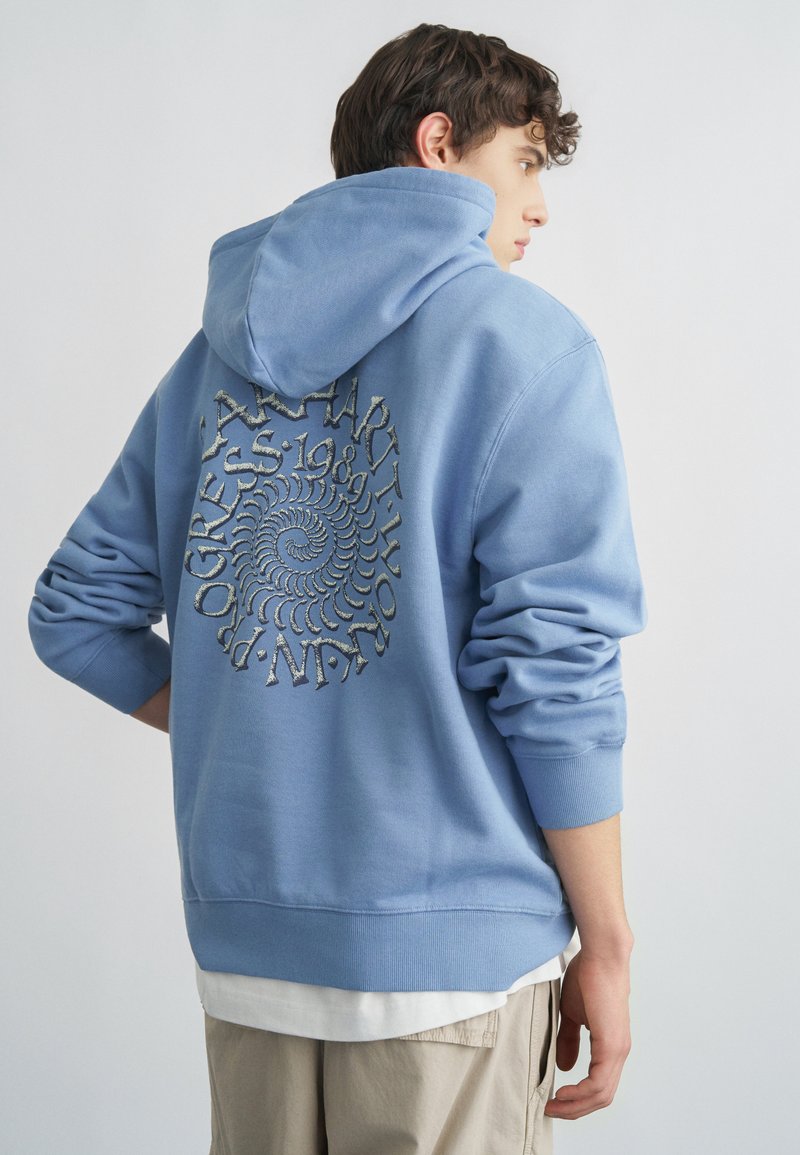 Noor mees seljaga, kandes helesinist kapuutsiga dressipluusi, millel on kapuuts peas ning seljal spiraalkujutis ja tekst, beeže püksid ja valge aluspluus.