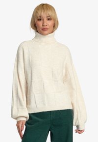 Kremowy, oversize'owy sweter z wysokim kołnierzem, teksturowanym wzorem w kwadraty i szerokimi rękawami. Połączony z ciemnozielonymi spodniami z kordurowej tkaniny.