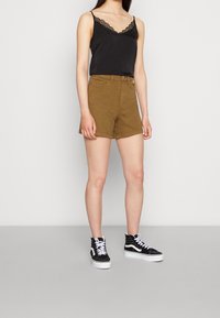 Camisole noire avec borde de dentelle, associée à un short marron clair. Le mannequin porte des baskets montantes noires avec des accents blancs. Fond neutre.