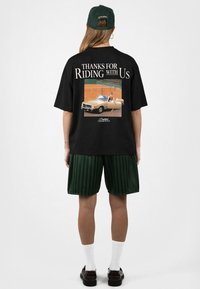 Personne portant un t-shirt noir avec le texte « Thanks for Riding With Us » et un graphisme de voiture vintage, portant un short vert, des chaussettes blanches et des chaussures noires.