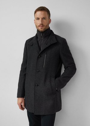Man draagt een donker textuurde wollen jas met knoopsluiting en een zwarte, hoogsluitende binnenlaag, staand tegen een lichtgrijze muur.