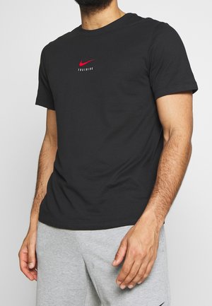 Czarna bawełniana koszulka z okrągłym dekoltem, krótkimi rękawami i luźnym krojem, z czerwonym logo Nike i napisem. Noszona z szarymi spodniami dresowymi.