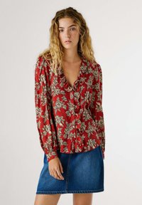 Blouse rouge à fleurs avec des manches longues et un col à volants, ornée de motifs de fleurs beiges. Associée à une jupe en jean.