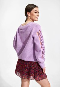 Paarse gebreide trui met open kantdetails op de mouwen, gecombineerd met een gelaagde mini-jurk in een bloemenprint in rode, roze en blauwe tinten.