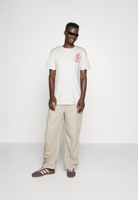 Camiseta blanca de mangas cortas con un diseño gráfico rojo en el pecho, combinada con pantalones beige de corte holgado y zapatos marrones con rayas blancas.