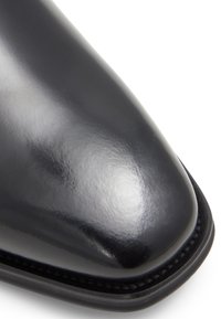 Černá kožená bota s lesklým povrchem, špičatou špičkou a kontrastním šitím podél podrážky. Hladká textura, elegantní design a žádné viditelné kovové prvky.