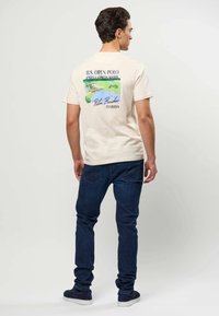 Mand iført beige U.S. Open Polo Championship T-shirt med Palm Beaches Florida grafik, mørkeblå jeans og marineblå sneakers, vendt væk.