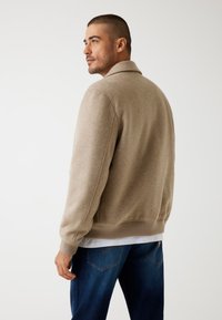 Beige trui van een zachte, gestructureerde stof met een kraagontwerp en geribde manchetten. Draagt over een wit shirt en gecombineerd met blauwe jeans.