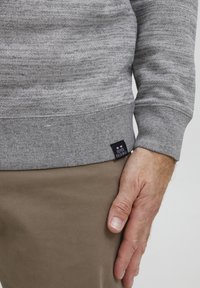 Gray sweater with a textured finish, ribbed cuffs, and hem. Features a small, branded label sewn at the bottom left. Paired with beige pants.