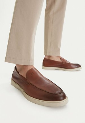 Braune Leder-Slipper mit hellen Sohlen, getragen zu beigen Hosen, seitlich und schräg von vorne gezeigt.