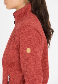 Röd fleecjacka med hög krage, dragkedjeficka på sidan, texturerad tyg och en liten ljusbrun logotyp på övre ärmen.