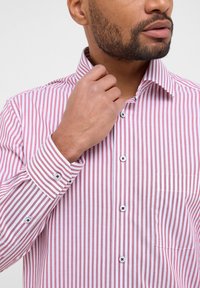 Camicia a righe rosse e bianche con colletto, maniche lunghe e taschino sul petto. Presenta bottoni solidi con un design contrastante.