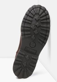 Bota de cuero marrón con una suela de goma negra robusta, que presenta profundas tracciones y un grabado de logo. Diseño duradero para uso al aire libre.