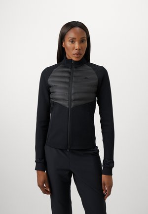 Kvinna iförd en svart vadderad och ribbad zip-up jacka med matchande svarta byxor, stående mot en enkel ljus bakgrund.