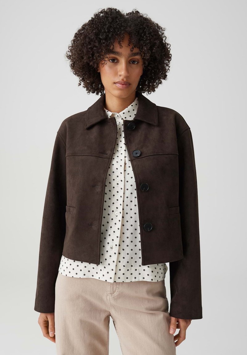 Woman with curly hair wearing a dark brown buttoned jacket over a white polka dot shirt and light beige pants, standing against a plain background.