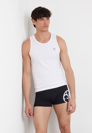 Débardeur en coton blanc avec un petit logo noir, associé à un short de boxeur noir arborant un emblème stylisé blanc. Se tenant contre un mur blanc.