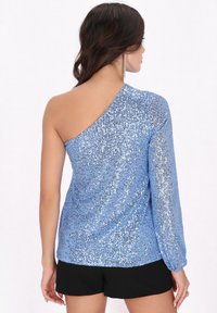 Abito asimmetrico in tessuto blu chiaro con paillettes, caratterizzato da una manica lunga, superficie texturizzata e scollatura profonda sulla schiena. Indossato con pantaloni corti neri.