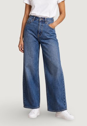 Personne portant un jean bleu ample taille haute, des baskets blanches et un haut blanc à manches courtes, une main dans la poche.