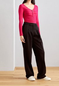 Top aderente rosa a coste con chiusura a zip e pantaloni neri a vita alta plissettati. Il modello indossa sneakers bianche. Texture morbide e colori vivaci.