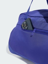 Μαύρο smartphone με τριπλή κάμερα, εν μέρει μέσα στην μπροστινή τσέπη με φερμουάρ μιας μπλε υφασμάτινης τσάντας duffel με χερούλι.