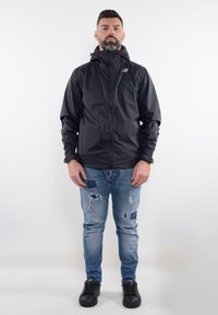 Black waterproof jacket with a zip front and hood, featuring a logo on the chest. Worn with light-blue distressed jeans and black shoes.