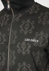 Chaqueta negra con cremallera y un patrón damasco tonal. En la parte frontal presenta el texto en blanco "LES DEUX". La tela es suave, con un cuello alto y una cremallera metálica.