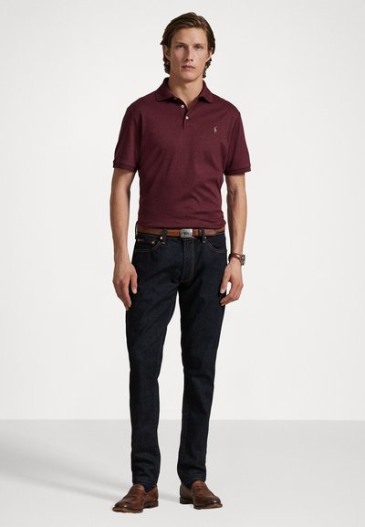 Cămașă polo roșie închis cu guler, asociată cu blugi negri. Curea și pantofi maro completează ținuta. Design simplu și textură netedă.