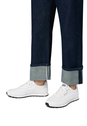 Sneakers atletici bianchi con suole nere; linguetta imbottita, design con lacci e logo laterale. I jeans hanno orli arrotolati con denim in contrasto.